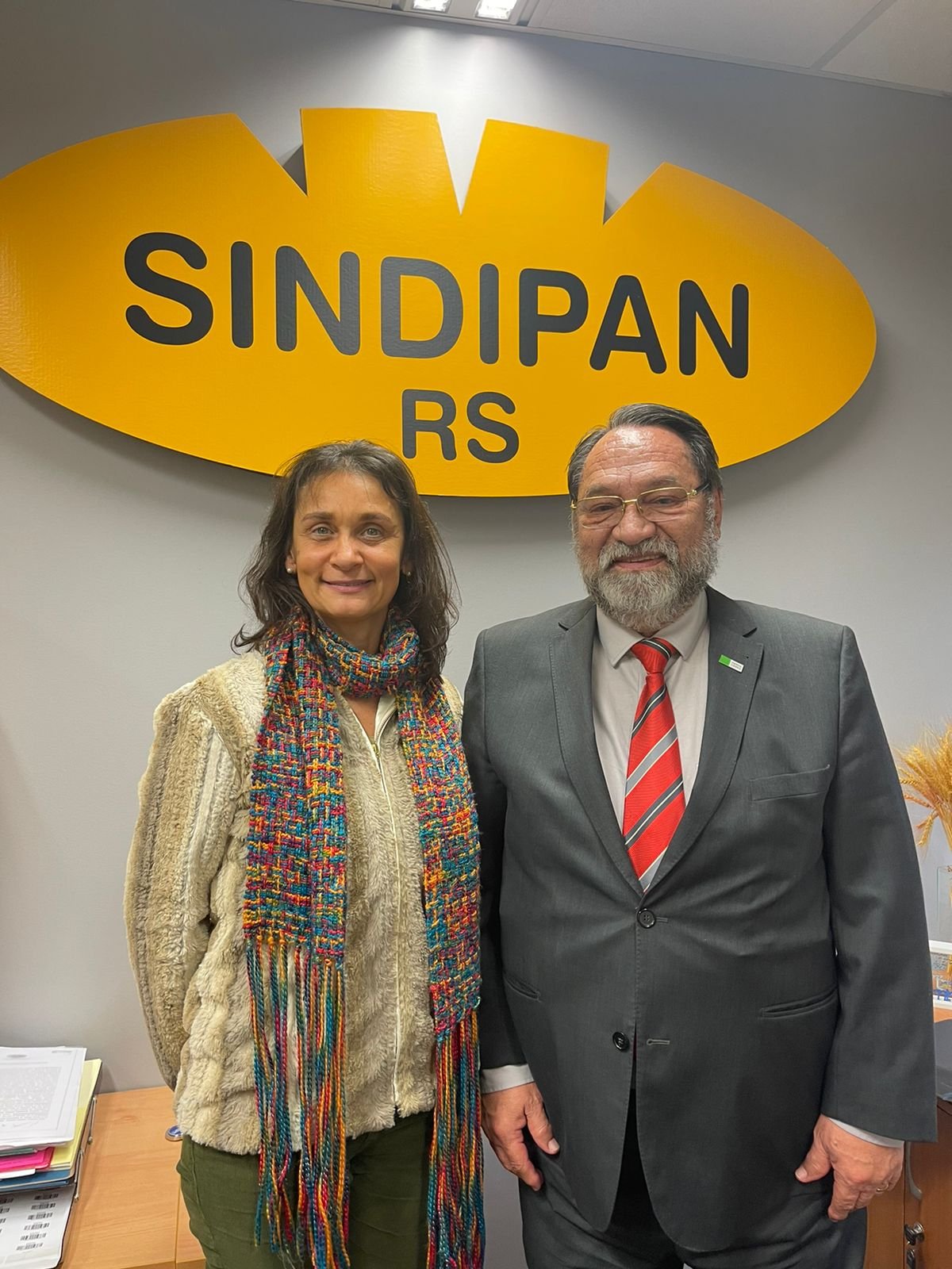 Um homem de barba e uma mulher à frente de uma parede branca com o símbolo do Sindipan RS em amarelo